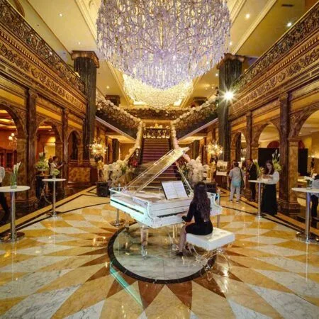 A pianist performs on a grand piano under a large chandelier in a luxurious hall.